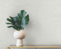A vase containing large green monstera leaves sits on a narrow table against a wall with textured light gray Winn Faux Grasscloth Wallpaper.
