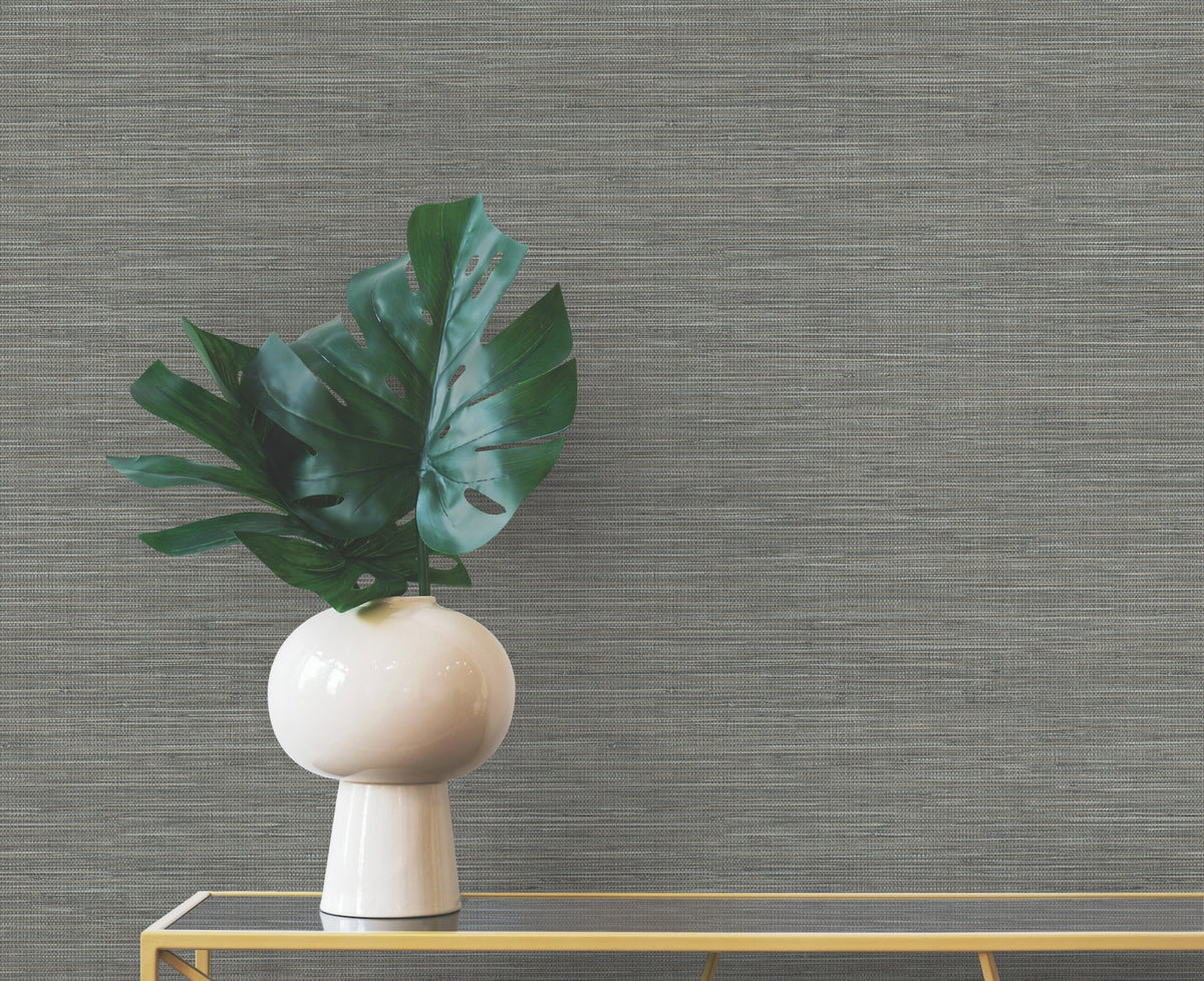 A modern white vase with a large monstera leaf sits on a gold console table against gray Winn Faux Grasscloth Wallpaper by York Wallcoverings.