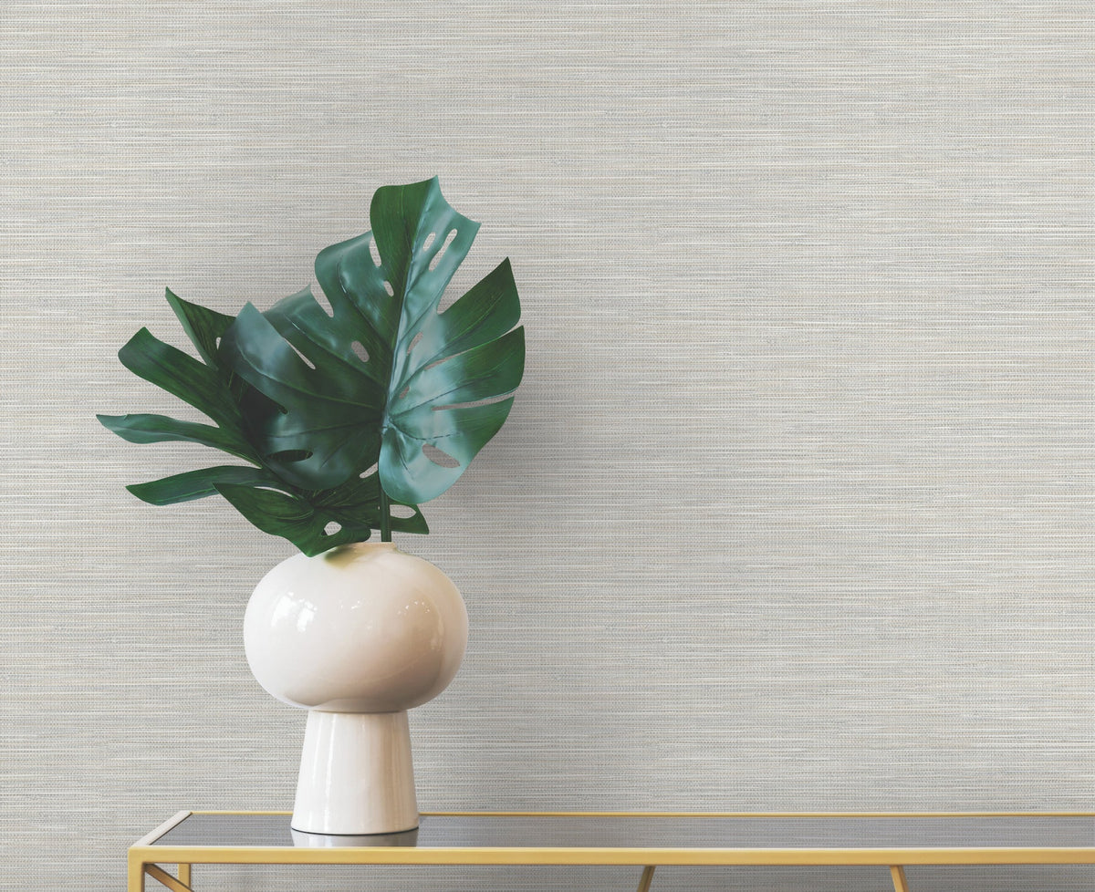 A white vase containing large monstera leaves sits on a narrow table against a wall covered in light gray Winn Faux Grasscloth Wallpaper by York Wallcoverings.