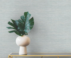 A large monstera leaf in a rounded white vase sits on a gold console table against light blue Winn Faux Grasscloth Wallpaper by York Wallcoverings.