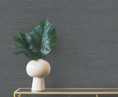 Winn Faux Grasscloth Wallpaper in dark gray provides a backdrop for a white vase containing green monstera leaves on a glass table.
