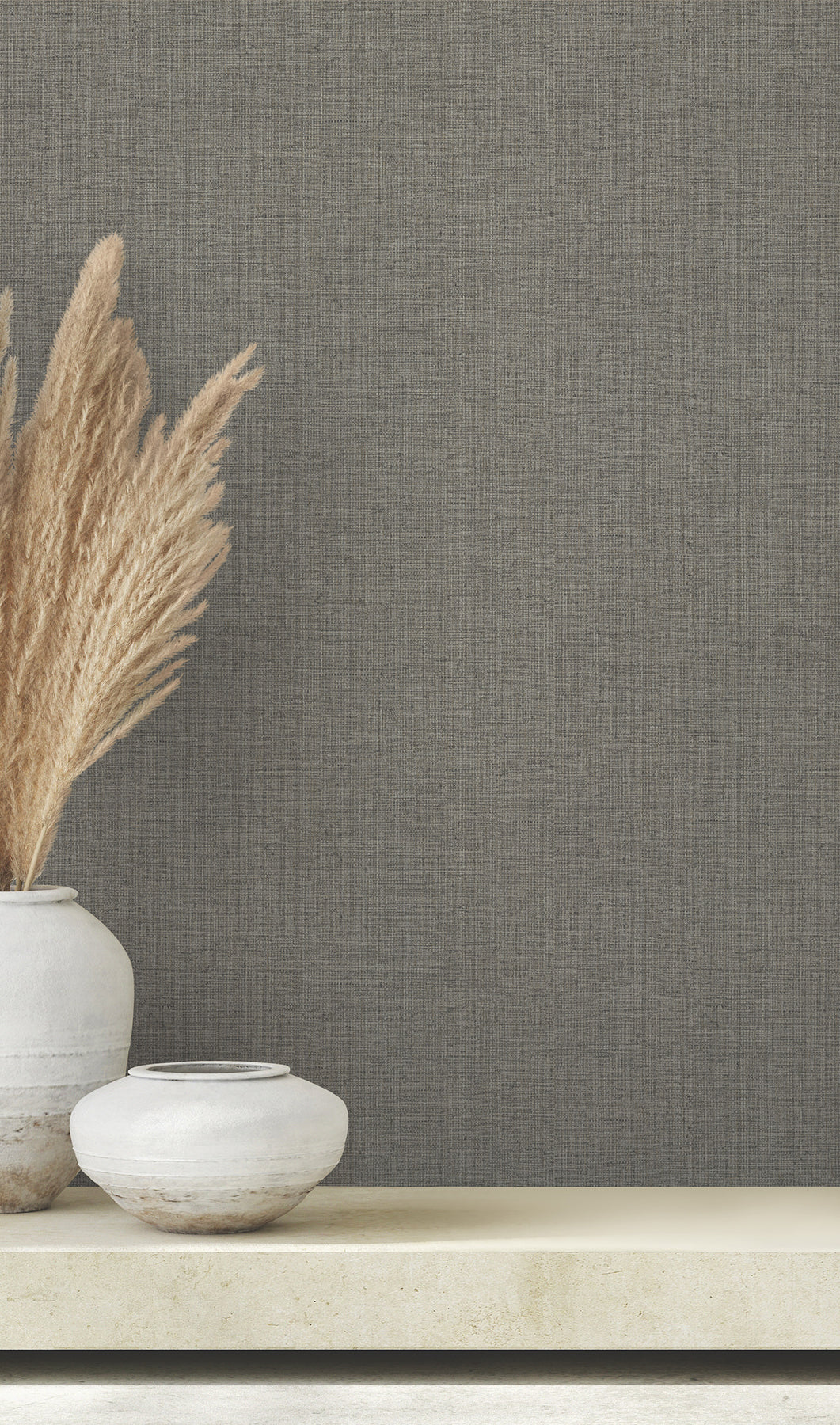 Clarkson Woven Wallpaper in dark gray with a subtle woven texture, shown behind a shelf with two ceramic vases and tall dried grass.