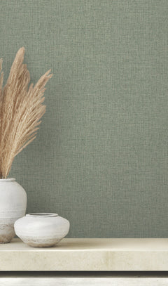 Clarkson Woven Wallpaper in sage green with a woven fabric texture, shown behind a shelf with two ceramic vases and pampas grass.