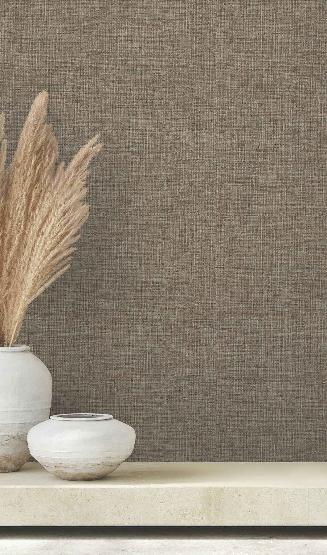 Two ceramic vases with dried grasses set against brown Clarkson Woven Wallpaper by York Wallcoverings.