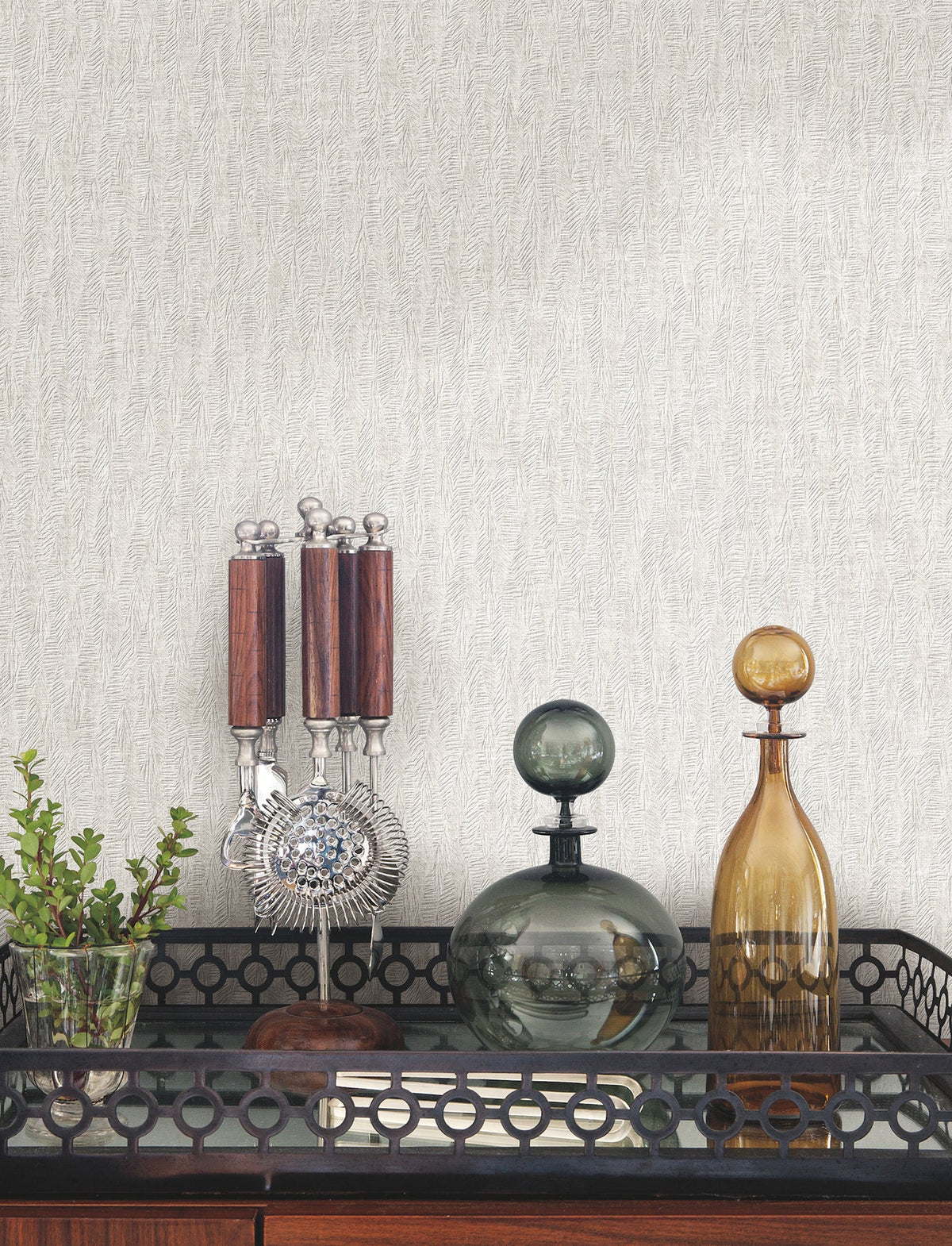 Bryson Feathered Texture Wallpaper in light gray with a fine, feathered texture displayed behind a bar cart holding a glass plant vase, metal bar tools, and two glass decanters.