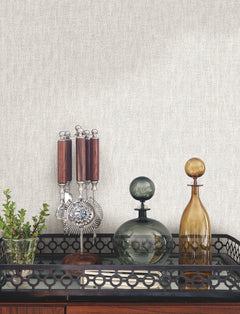 Bryson Feathered Texture Wallpaper in light gray with a fine, feathered texture displayed behind a bar cart holding a glass plant vase, metal bar tools, and two glass decanters.