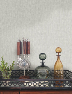 Bryson Feathered Texture Wallpaper in light gray with a fine, vertical feathered texture behind a black metal bar cart holding a bar tool set, glass decanters, and a small potted plant.