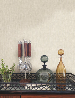A bar cart with a glass plant vase, a stand of bar tools, and two glass decanters set against light-colored Bryson Feathered Texture Wallpaper by York Wallcoverings with a subtle vertical texture.