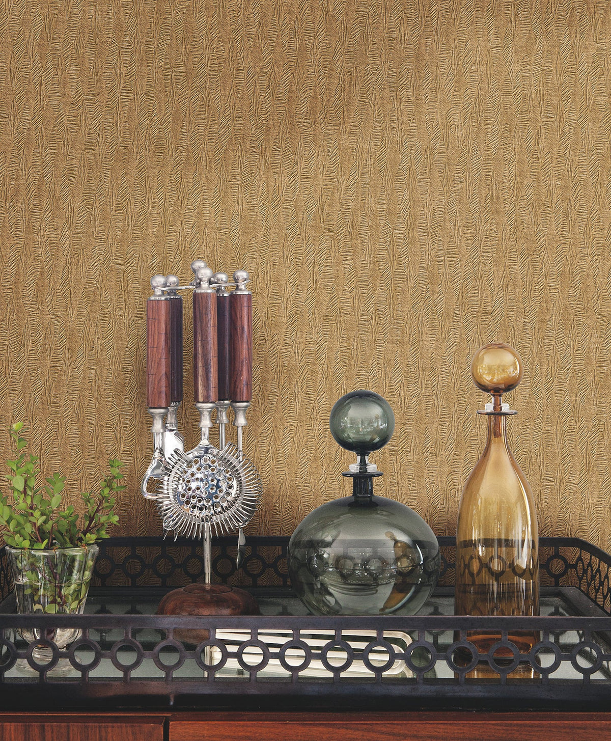 Gold-toned Bryson Feathered Texture Wallpaper with a subtle feathered texture behind a bar cart containing a cocktail tool set and two glass decanters.