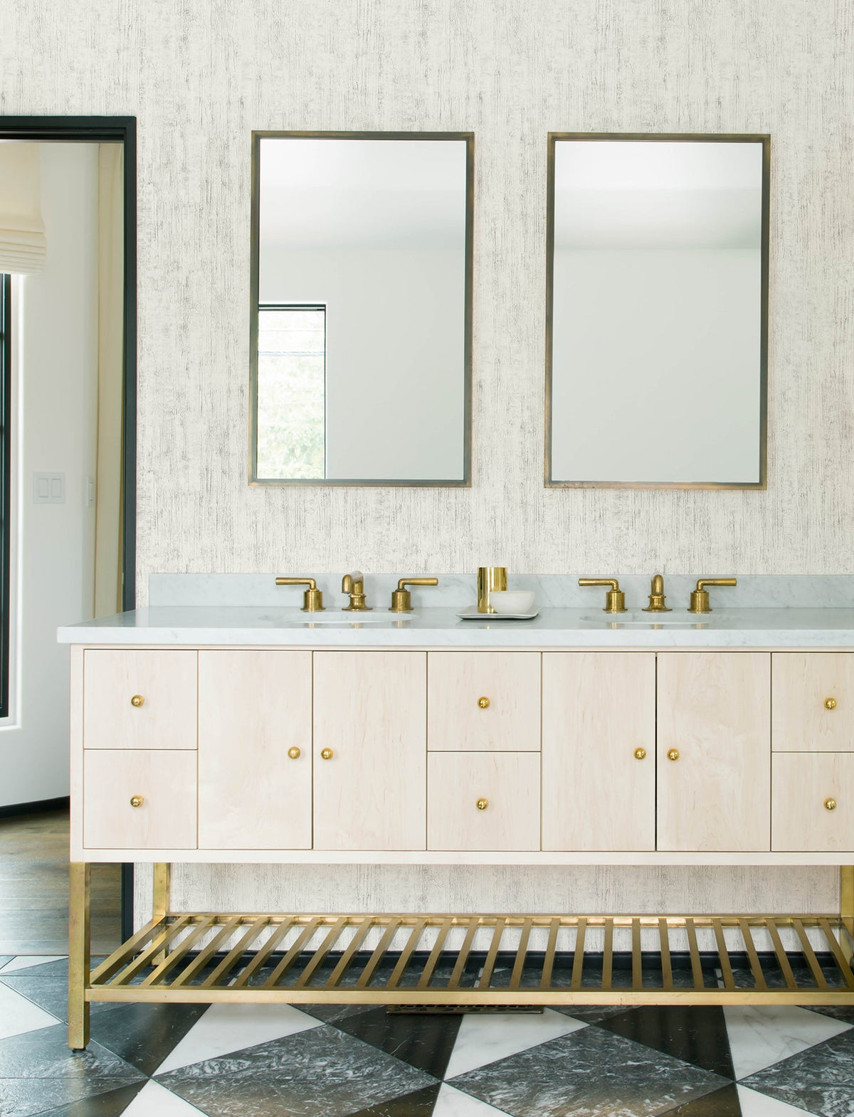 Modern bathroom featuring Chesapeake Ramona Distressed Texture Wallpaper by York Wallcoverings, double sinks with brass faucets, two mirrors, a light wood vanity with brass accents, and black-and-white checkered floor tiles.