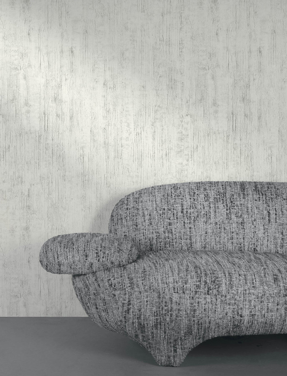A modern gray textured sofa sits in front of a wall covered with York Wallcoverings’ Chesapeake Ramona Distressed Texture Wallpaper in light gray. Only part of the sofa is visible, featuring its curved armrest and seat facing the viewer. The floor complements the scene with a matching shade of gray.