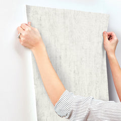 A person in a striped shirt aligns a piece of Chesapeake Ramona Distressed Texture Wallpaper by York Wallcoverings against a white wall, preparing to apply it.