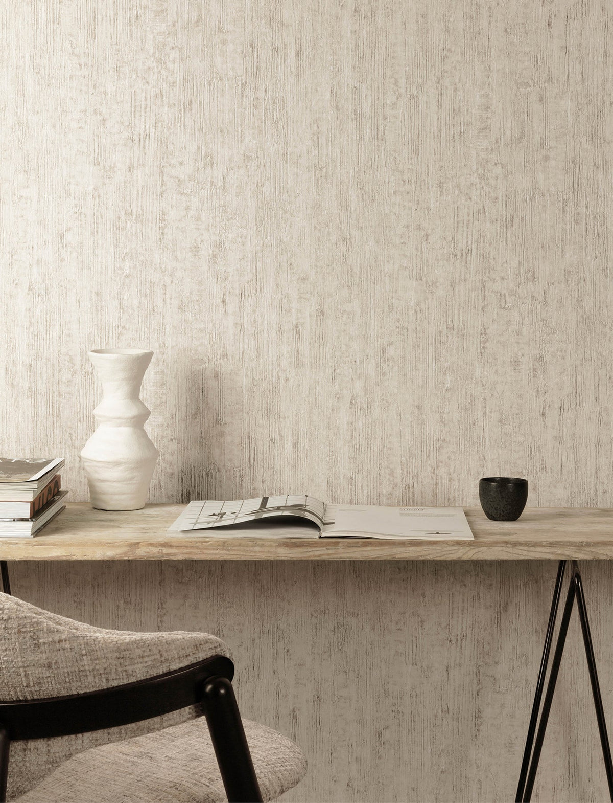 A minimal desk setup features a wall adorned with Chesapeake Ramona Distressed Texture Wallpaper by York Wallcoverings, a ceramic vase, stacked books, an open magazine, and a black cup. A light gray cushioned chair sits at the wooden desk.