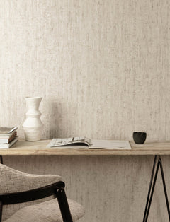 A minimal desk setup features a wall adorned with Chesapeake Ramona Distressed Texture Wallpaper by York Wallcoverings, a ceramic vase, stacked books, an open magazine, and a black cup. A light gray cushioned chair sits at the wooden desk.