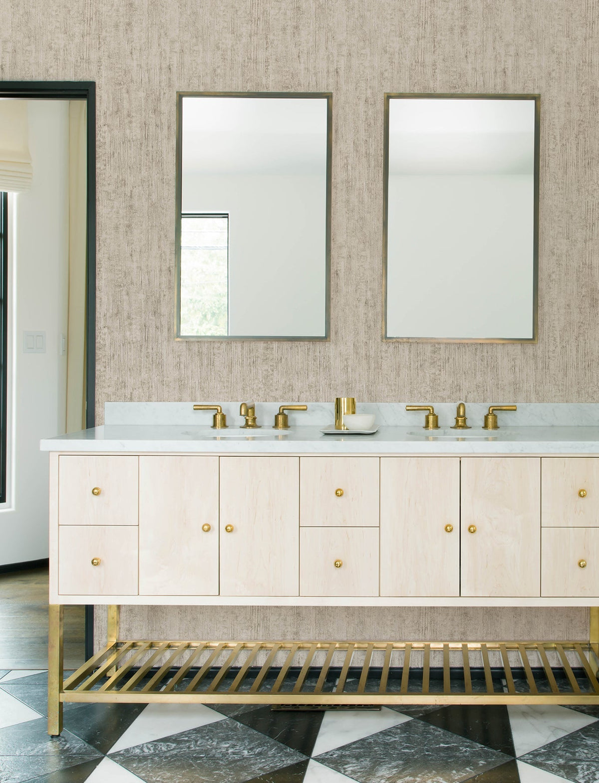 Modern bathroom with two rectangular mirrors above a double-sink vanity. The vanity features light wood cabinets, gold faucets, and an open slatted shelf. Black and white geometric floor tiles complement the neutral Chesapeake Ramona Distressed Texture Wallpaper by York Wallcoverings on the walls.