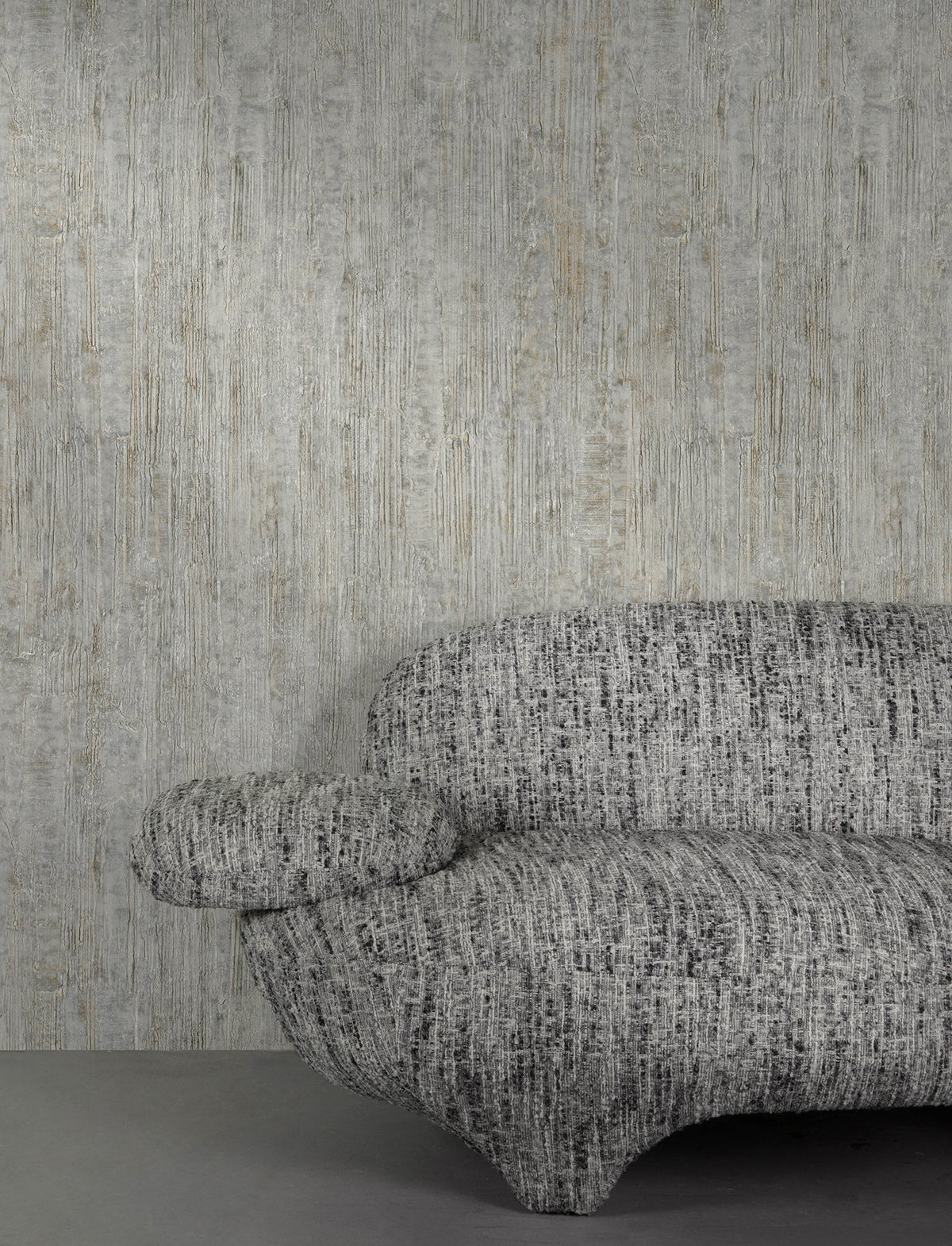 A modern, textured gray sofa with rounded shapes sits against a wall covered in York Wallcoverings’ Chesapeake Ramona Distressed Texture Wallpaper. Only the right half of the sofa is visible. The floor is a smooth, solid gray.