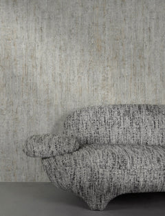A modern, textured gray sofa with rounded shapes sits against a wall covered in York Wallcoverings’ Chesapeake Ramona Distressed Texture Wallpaper. Only the right half of the sofa is visible. The floor is a smooth, solid gray.