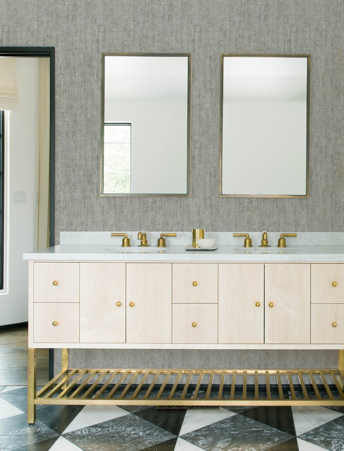 Modern bathroom with two mirrors, gold faucets, and a light wood vanity with gold accents. The floor features a black and white geometric tile pattern, while the walls are adorned with Chesapeake Ramona Distressed Texture Wallpaper by York Wallcoverings in a neutral tone.