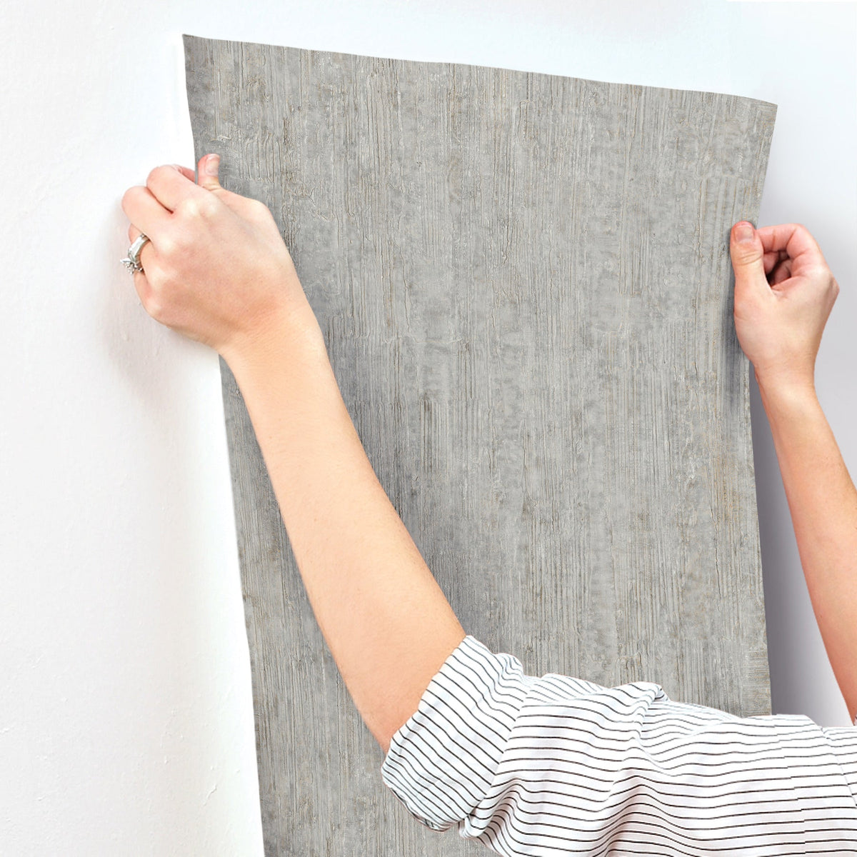 Wearing a striped shirt, a person applies Chesapeake Ramona Distressed Texture Wallpaper by York Wallcoverings onto a white wall, using both hands to smooth the textured gray wallpaper into place.
