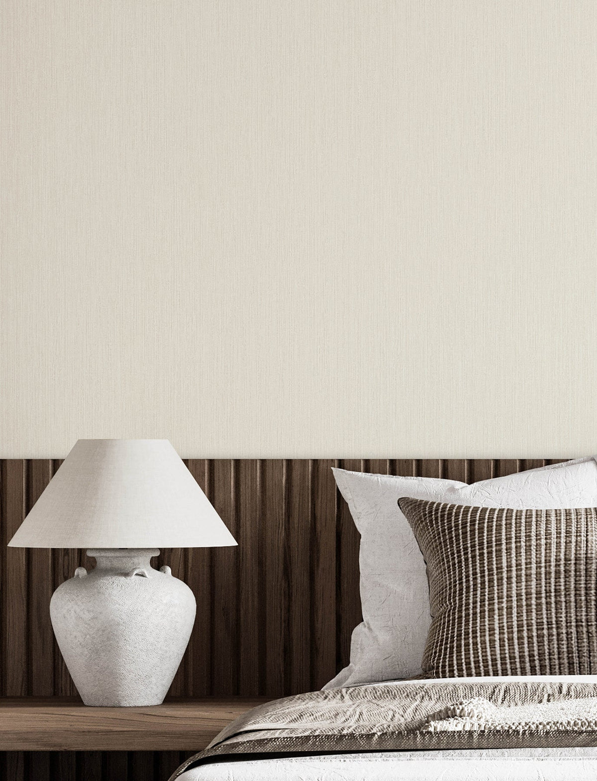 A minimalist bedroom scene featuring a white ceramic lamp on a wooden nightstand, brown-striped throw pillows, and Chesapeake Elijah Texture Wallpaper by York Wallcoverings in beige as the background. Only part of the bed and nightstand are visible.