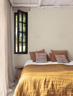 A cozy bedroom features the Chesapeake Elijah Texture Wallpaper by York Wallcoverings, complemented by mustard yellow bedding, decorative pillows, a rustic wooden bench, and beige curtains. A black-framed window reveals lush greenery outside, while white ceiling beams complete the serene, minimalist decor.
