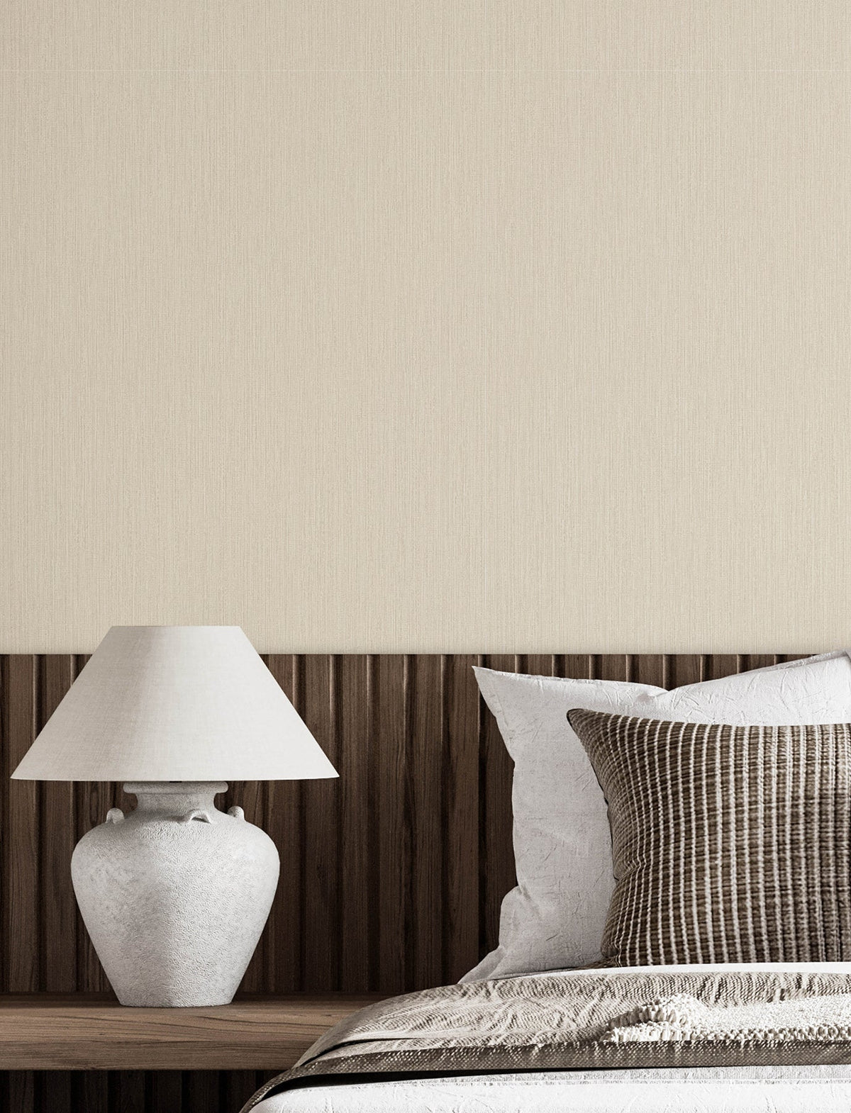 A white table lamp sits on a wooden headboard next to a bed with white and brown striped pillows, with Chesapeake Elijah Texture Wallpaper by York Wallcoverings covering the wall in the background.