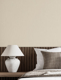 A white table lamp sits on a wooden headboard next to a bed with white and brown striped pillows, with Chesapeake Elijah Texture Wallpaper by York Wallcoverings covering the wall in the background.