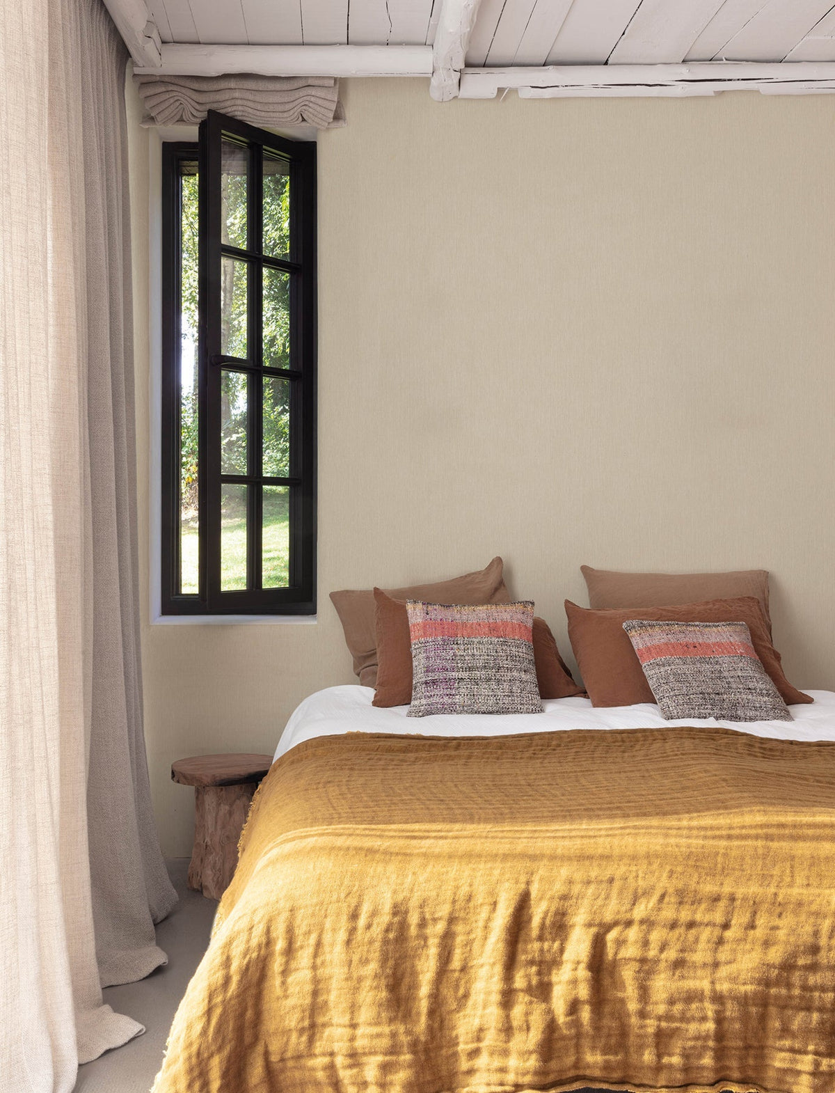 Cozy bedroom featuring Chesapeake Elijah Texture Wallpaper by York Wallcoverings, a mustard-colored bedspread, brown and patterned pillows, a rustic wooden bench, light beige curtains, and a black-framed window with greenery outside.