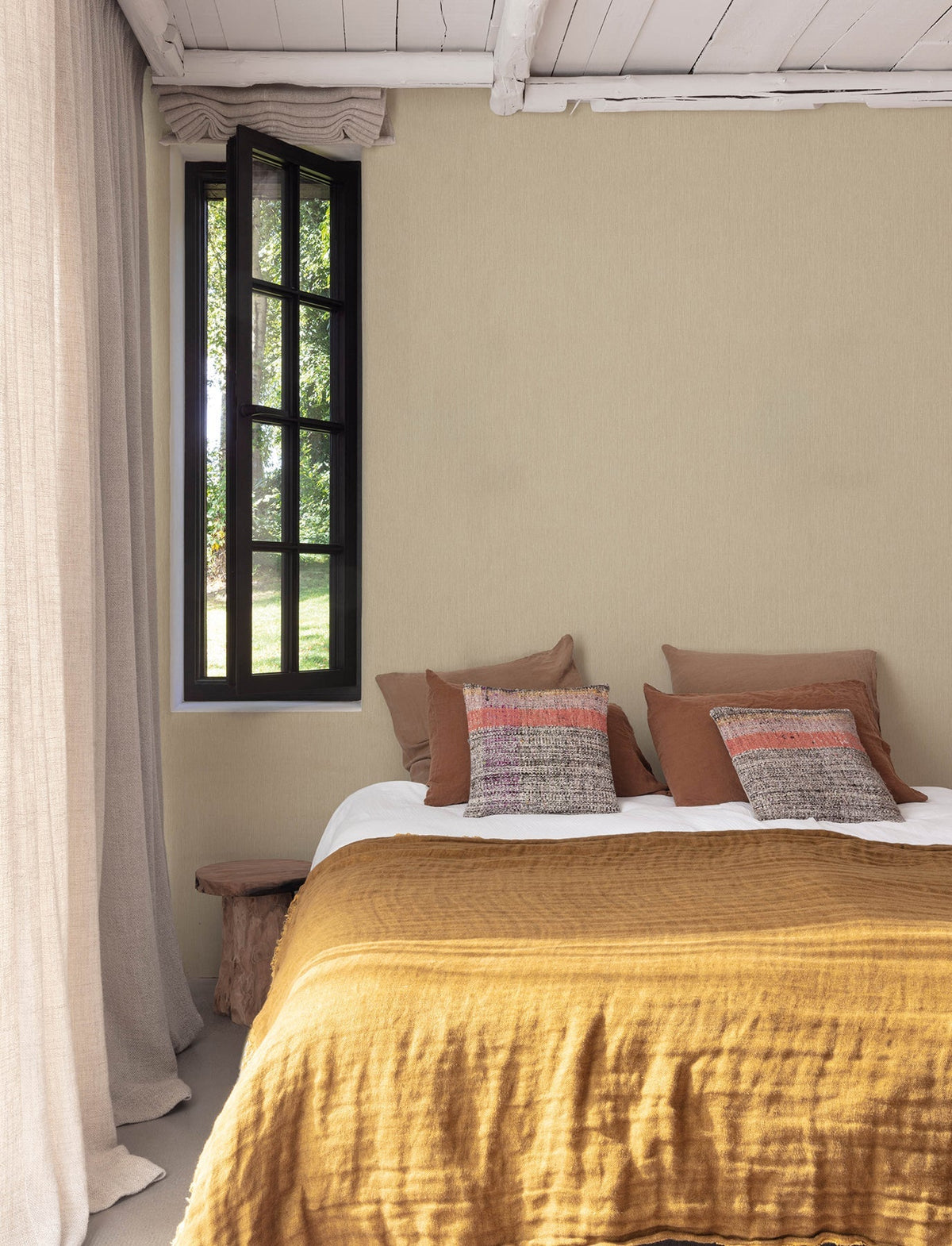 A cozy bedroom features a mustard-yellow bedspread, earth-toned pillows, and a rustic wood bench, with walls adorned in York Wallcoverings’ Chesapeake Elijah Texture Wallpaper. A black-framed window reveals greenery outside, and light curtains hang to the left.
