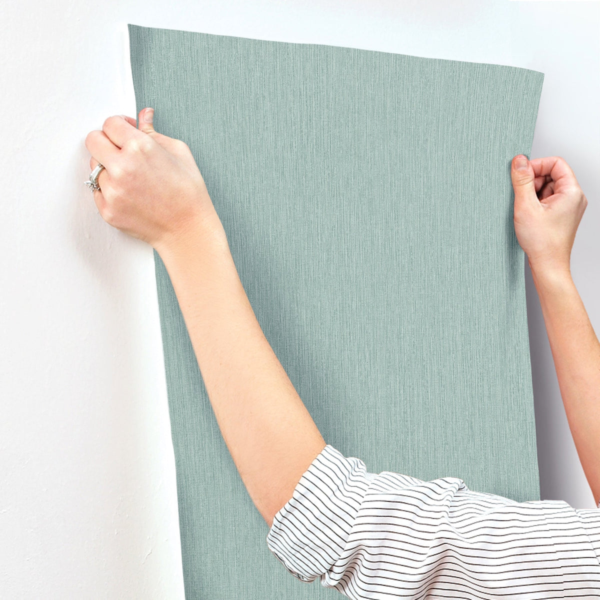 A person with a striped sleeve applies York Wallcoverings Chesapeake Elijah Texture Wallpaper in light blue onto a white wall, smoothing it with both hands.