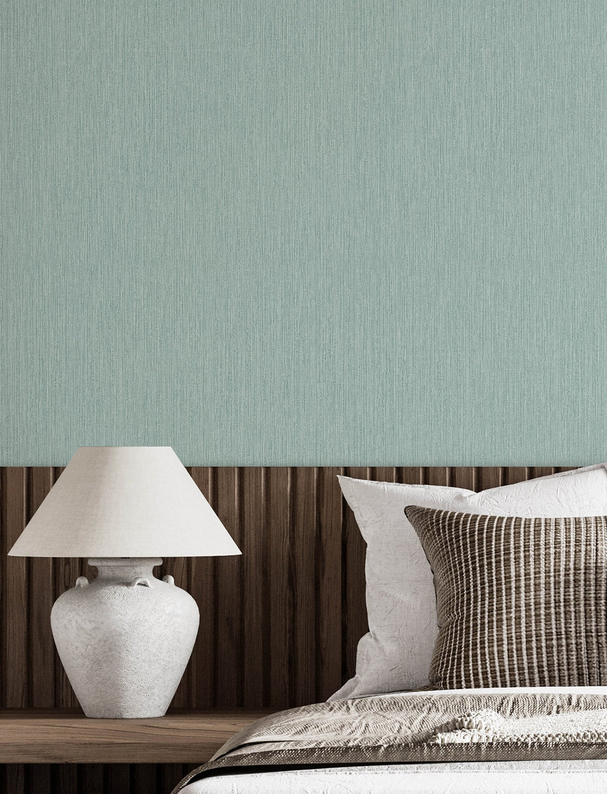 A neatly made bed with white and striped pillows sits next to a wooden headboard and a white ceramic lamp, set against a light green wall adorned with Chesapeake Elijah Texture Wallpaper by York Wallcoverings.