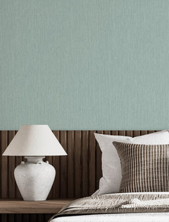 A neatly made bed with white and striped pillows sits next to a wooden headboard and a white ceramic lamp, set against a light green wall adorned with Chesapeake Elijah Texture Wallpaper by York Wallcoverings.