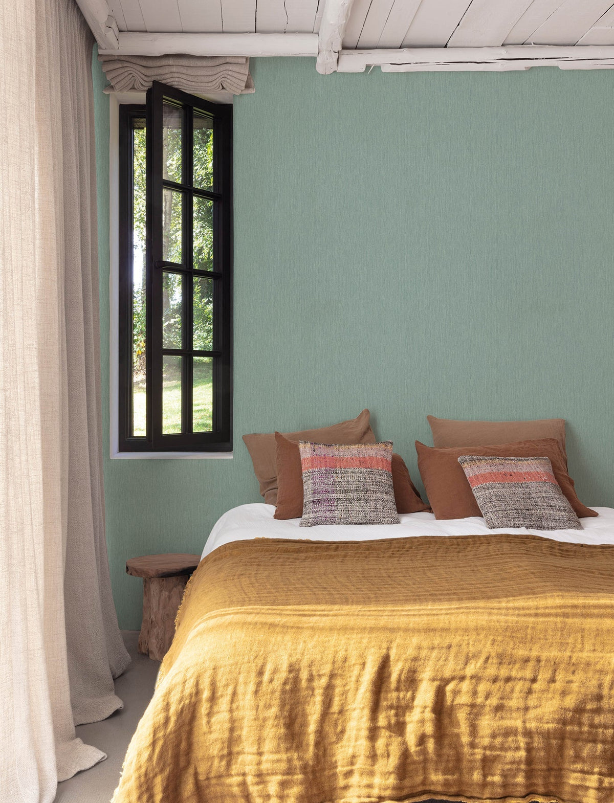 A cozy bedroom featuring a mustard yellow blanket, brown and patterned pillows, a wooden side table, sheer curtains, and a black-framed window overlooking greenery, with the walls adorned in Chesapeake Elijah Texture Wallpaper by York Wallcoverings.