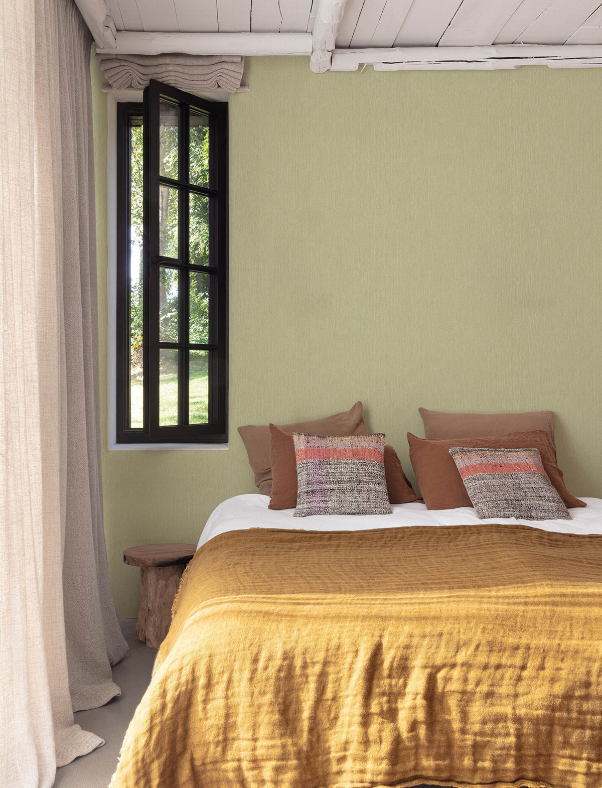A cozy bedroom featuring a mustard yellow bedspread, brown and patterned pillows, light green Chesapeake Elijah Texture Wallpaper by York Wallcoverings, a rustic wooden bench, sheer beige curtains, and a black-framed window with views of greenery outside.