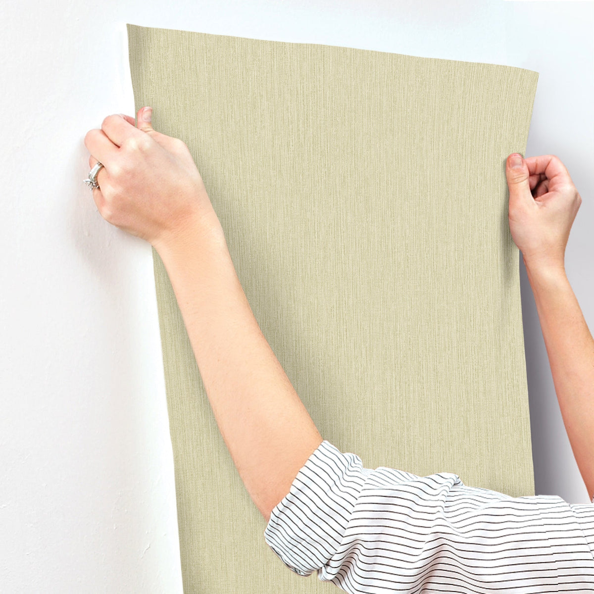 A person in a striped shirt applies Chesapeake Elijah Texture Wallpaper by York Wallcoverings to a white wall, smoothing it with both hands.