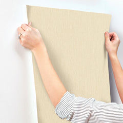 A person wearing a striped shirt is applying Chesapeake Elijah Texture Wallpaper by York Wallcoverings to a white wall, smoothing it out with both hands.