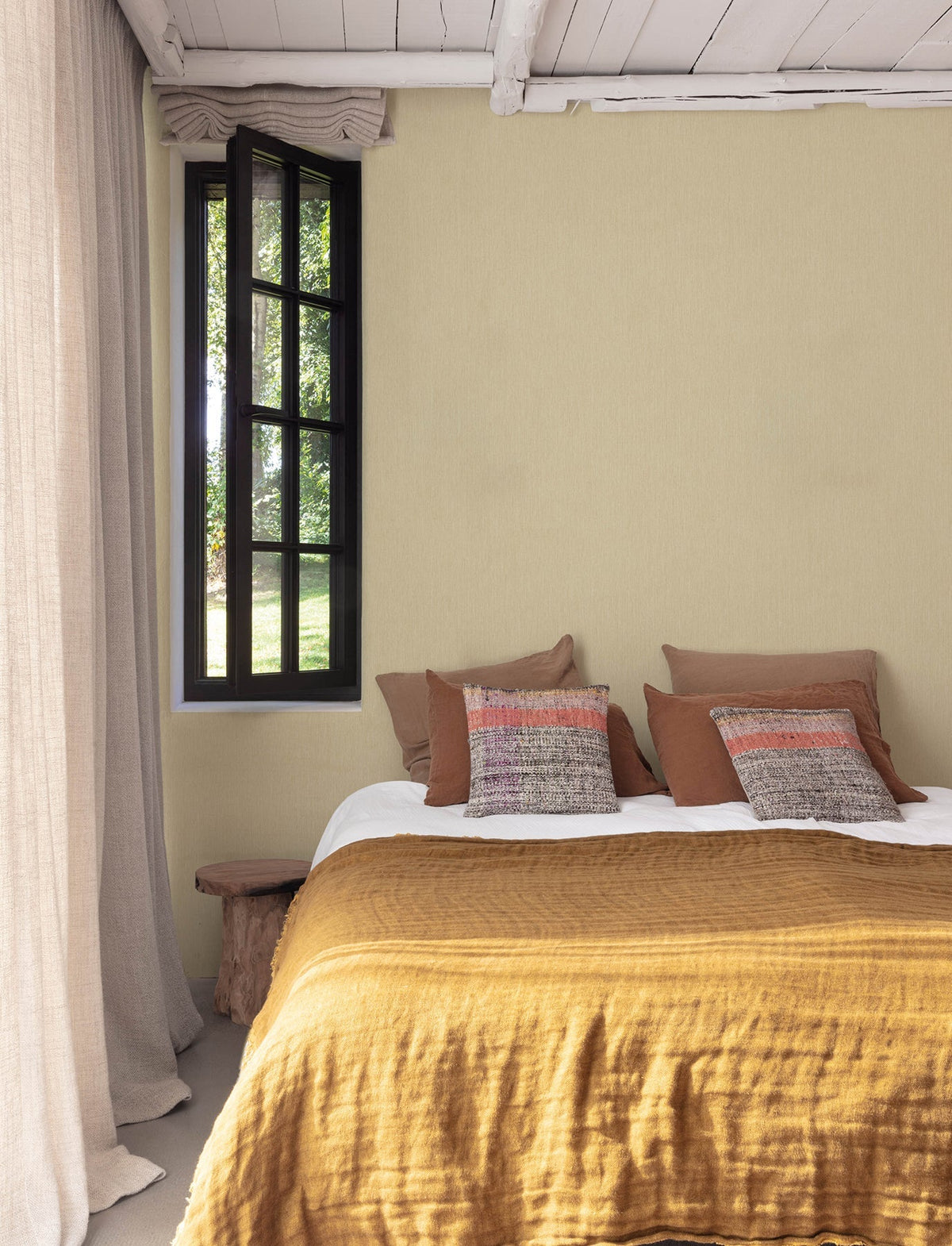 A cozy bedroom featuring Chesapeake Elijah Texture Wallpaper by York Wallcoverings, a mustard yellow bedspread, brown and patterned pillows, a rustic wooden bench, a window with black trim, and beige curtains. Light from the window brightens the space.