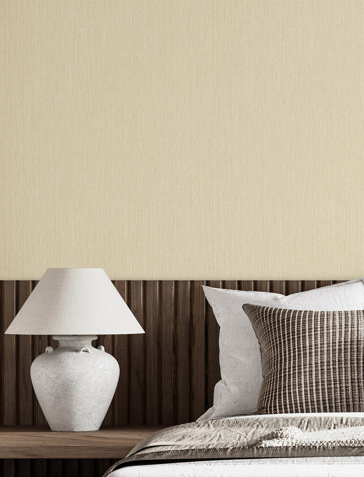 A white ceramic lamp sits on a wooden nightstand next to a bed with white and striped pillows, a wooden headboard, and walls adorned with York Wallcoverings’ Chesapeake Elijah Texture Wallpaper in a minimalist bedroom.