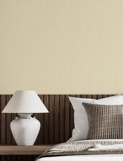 A white ceramic lamp sits on a wooden nightstand next to a bed with white and striped pillows, a wooden headboard, and walls adorned with York Wallcoverings’ Chesapeake Elijah Texture Wallpaper in a minimalist bedroom.