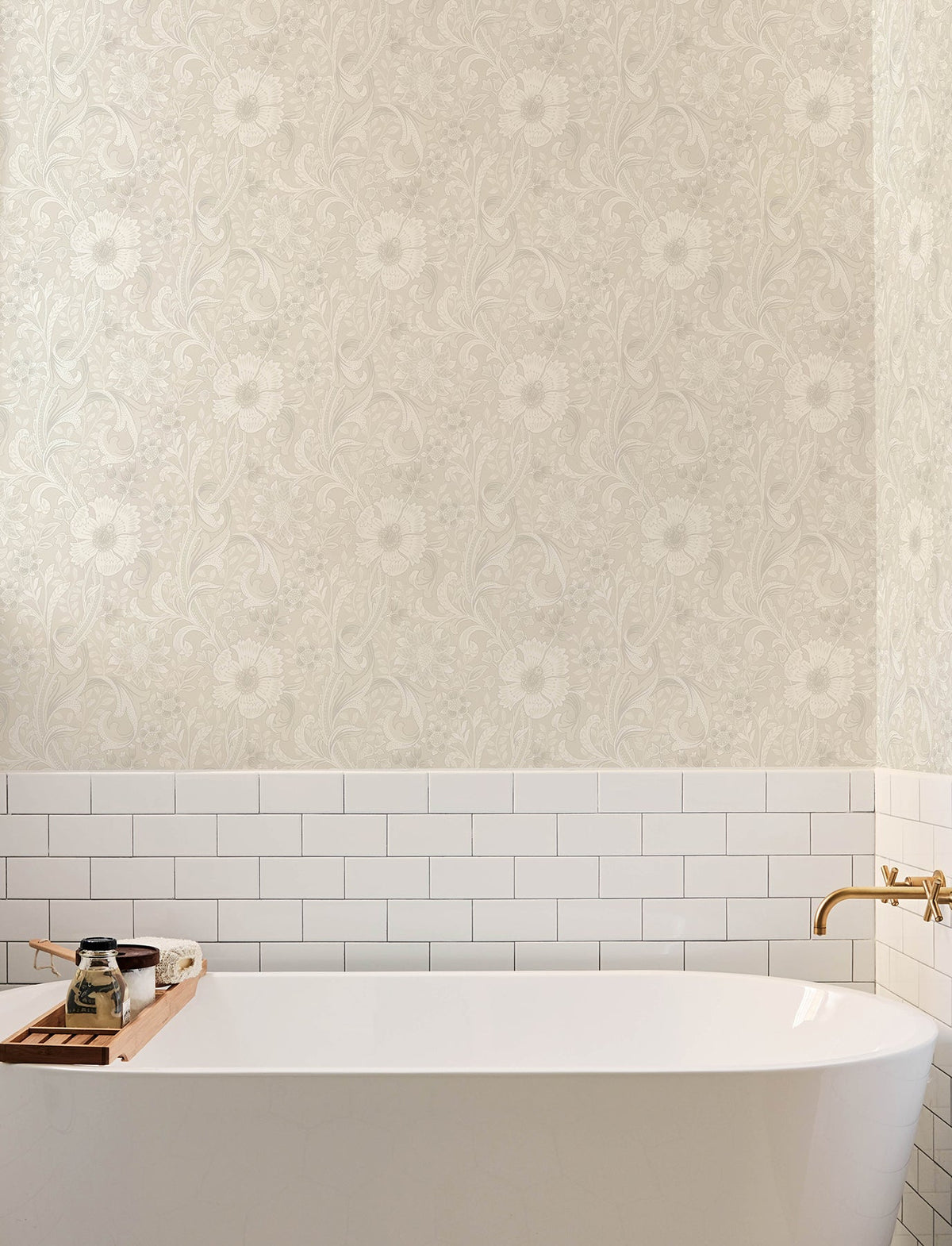 A white bathtub with a wooden tray, gold faucet, white subway tile, and York Wallcoverings’ Chesapeake Liora Floral Trail Wallpaper in beige above.