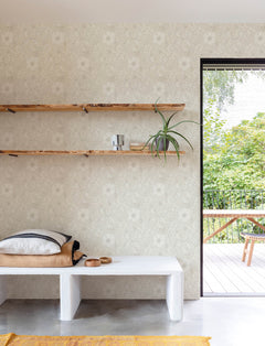 A minimalist room featuring York Wallcoverings’ Chesapeake Liora Floral Trail Wallpaper, with two wooden shelves displaying a plant and decor, a white bench topped with folded blankets and bowls, and an open door revealing a green patio.
