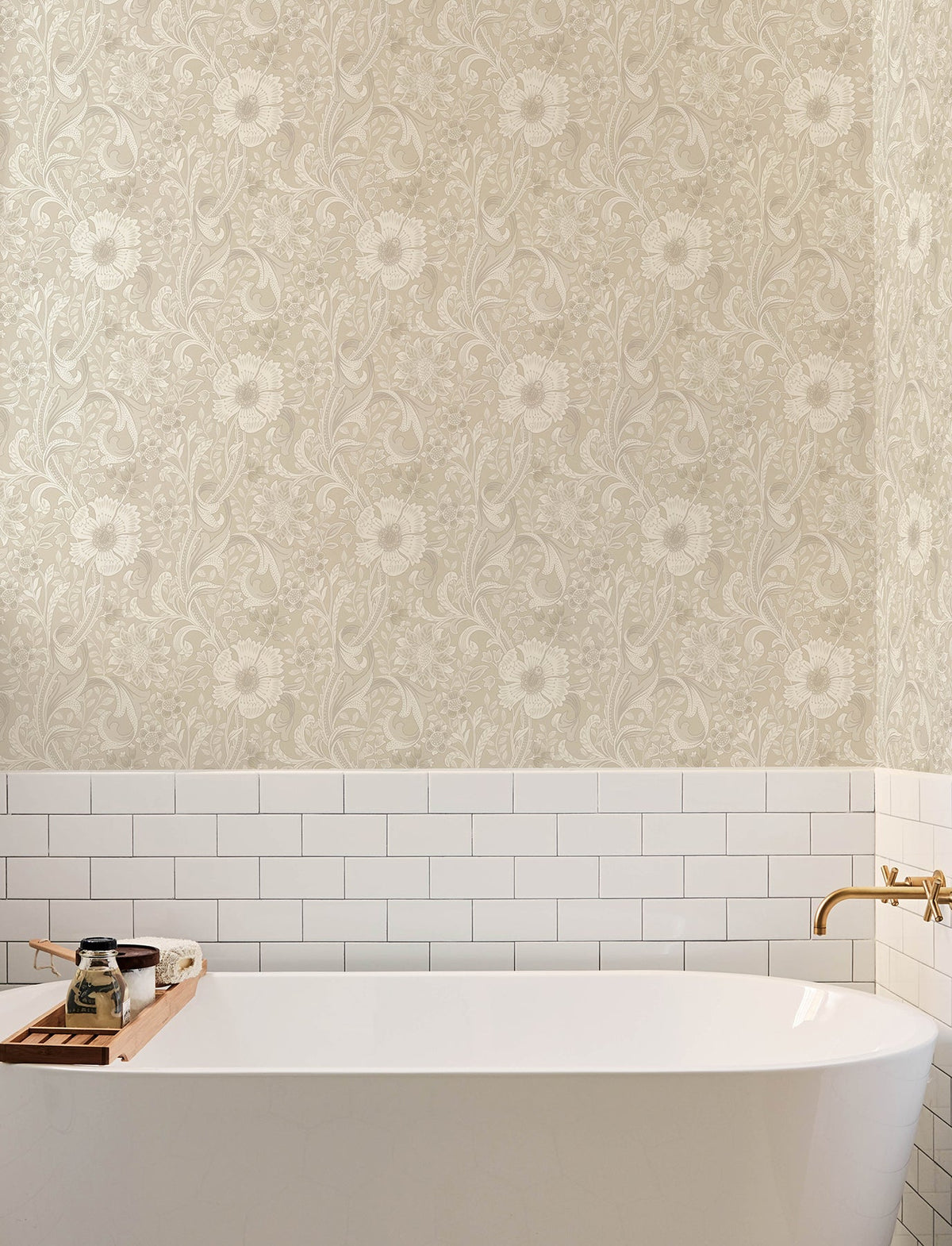 A white bathtub with a gold faucet sits against a wall adorned with York Wallcoverings’ Chesapeake Liora Floral Trail Wallpaper, featuring white subway tiles below. A wooden tray holds a candle, soap, and a small jar.
