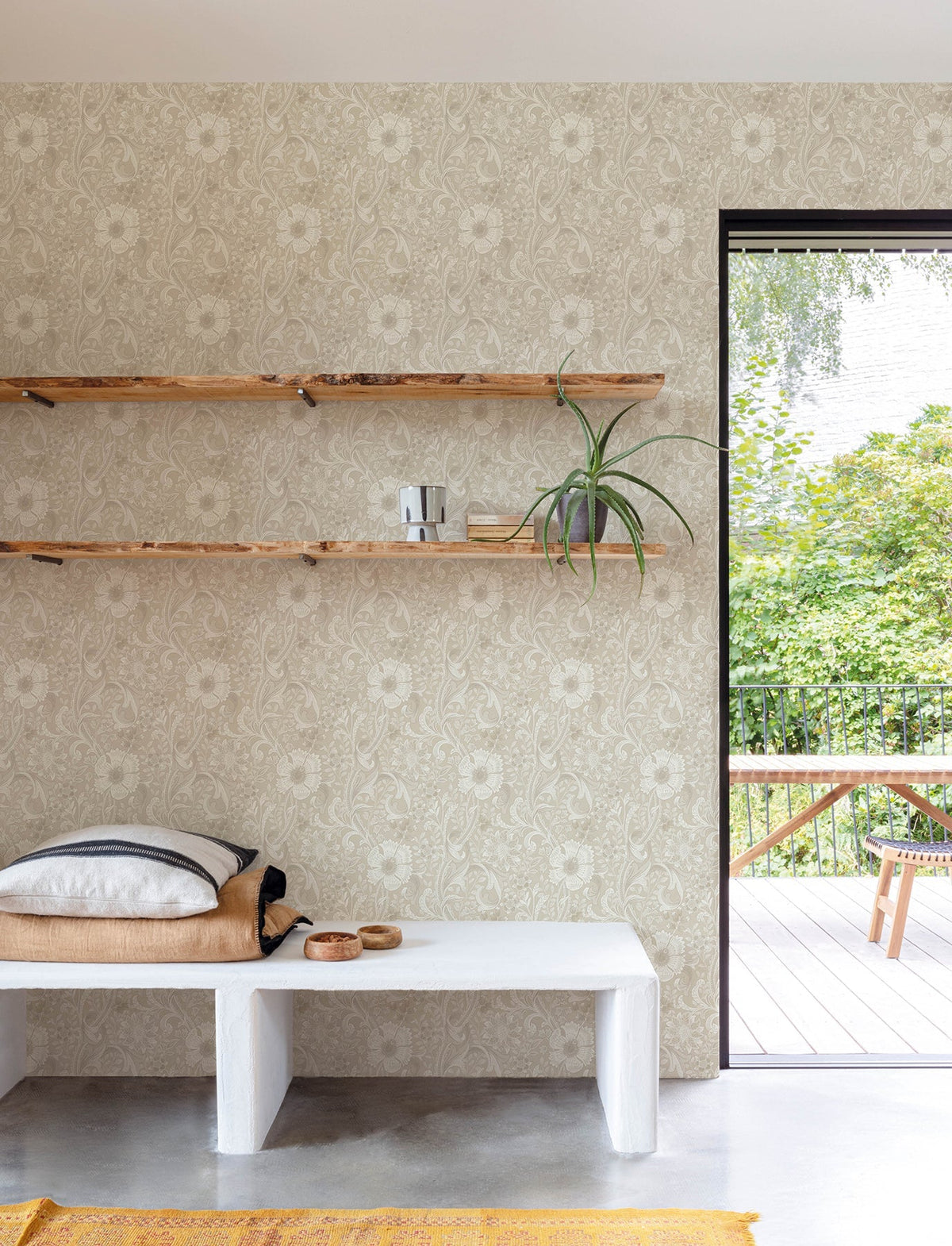 A minimalist room featuring York Wallcoverings’ Chesapeake Liora Floral Trail Wallpaper in beige, complemented by floating wooden shelves with a plant and decor, a white bench topped with folded linens, and a view of a green patio through a large open door.