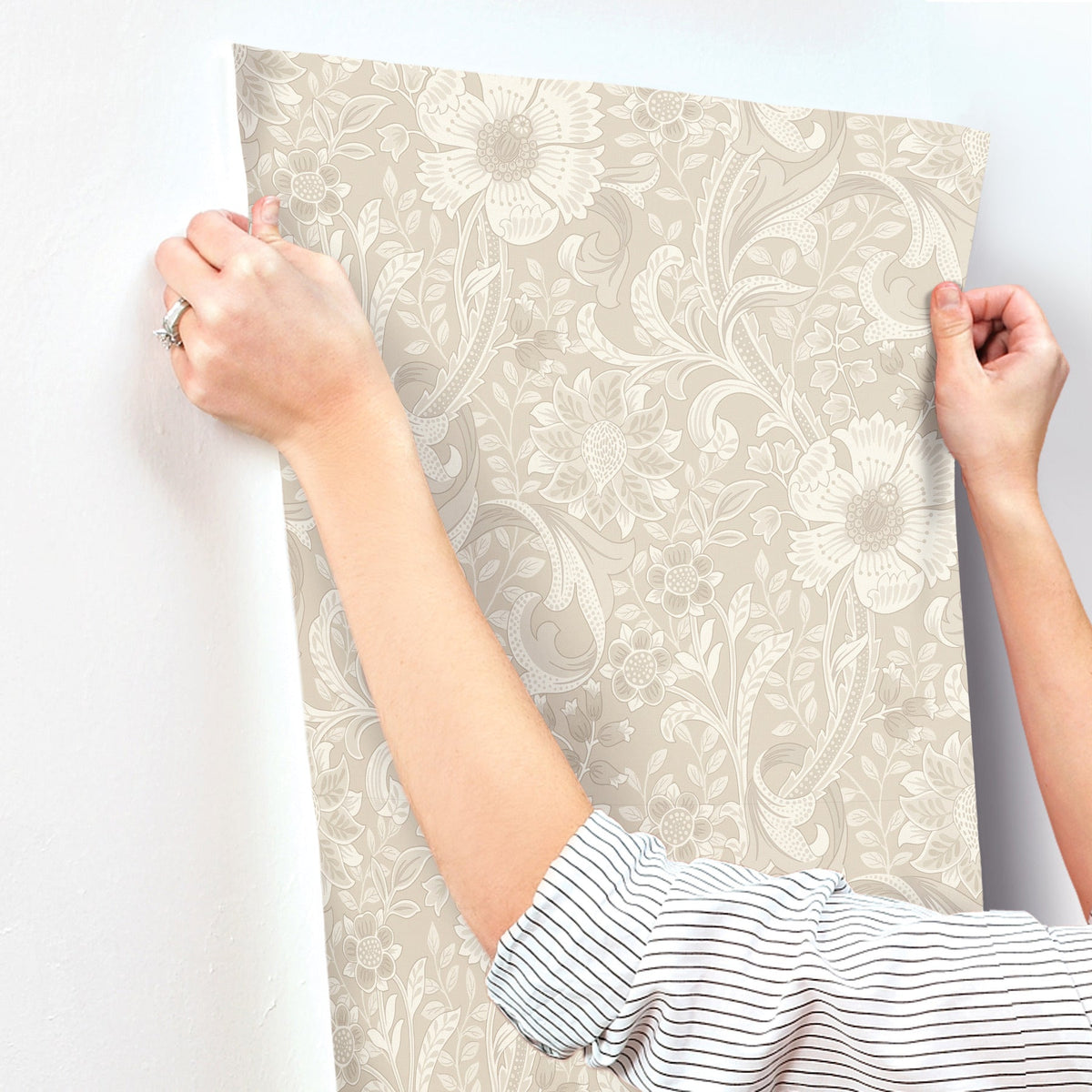 A person is applying York Wallcoverings Chesapeake Liora Floral Trail Wallpaper to a white wall, holding the wallpaper with both hands; only their arms and part of a striped shirt sleeve are visible.