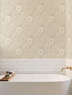 A white bathtub with a wooden tray holding soap and a candle is set against a wall adorned with Chesapeake Liora Floral Trail Wallpaper by York Wallcoverings and white subway tile. A gold faucet is mounted on the wall to the right.