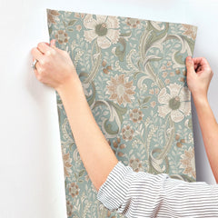 A person in a striped shirt is applying York Wallcoverings’ Chesapeake Liora Floral Trail Wallpaper, featuring green and beige floral patterns, to a white wall.