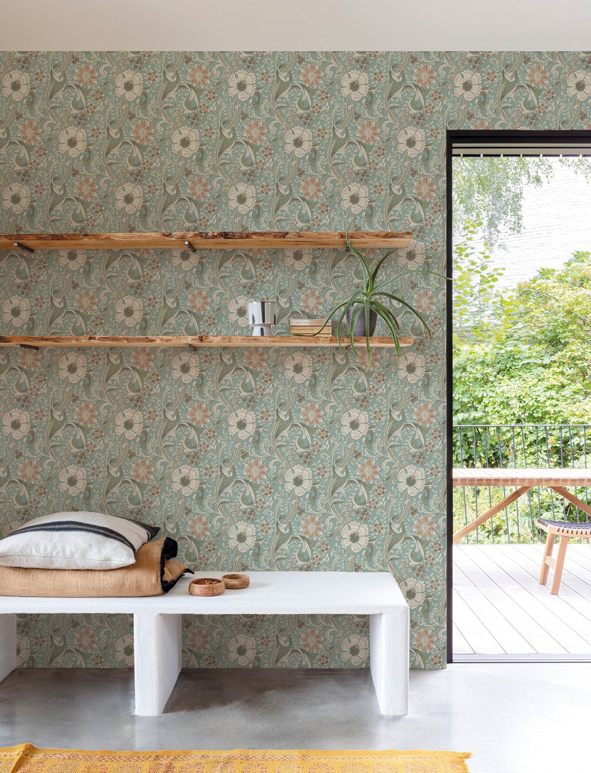 A cozy room featuring York Wallcoverings’ Chesapeake Liora Floral Trail Wallpaper, wooden shelves adorned with plants and decor, a cushioned bench, and a door open to a green outdoor patio with a table and benches.