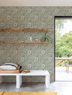 A room featuring Chesapeake Liora Floral Trail Wallpaper by York Wallcoverings, two wooden wall shelves holding books and a plant, a bench with pillows, a tray, and an open door leading to a green outdoor patio with a wooden table and bench.