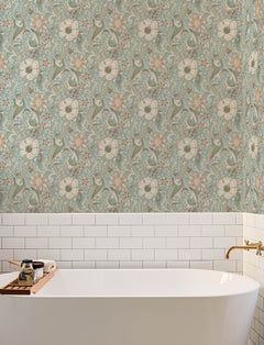 A white bathtub with gold fixtures sits against a wall adorned with York Wallcoverings’ Chesapeake Liora Floral Trail Wallpaper and white subway tiles. A wooden tray on the tub holds candles and bath accessories.