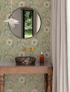 A round mirror above a marble sink, York Wallcoverings’ Chesapeake Liora Floral Trail Wallpaper, brass faucet, soap dispenser, and a hanging light fixture; beige curtain at the side and greenery visible through a window.